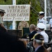 A military ceremony is being held at a Vietnam Veterans Association Northern Territory Inc. event. A female naval officer in white dress uniform with medals is speaking at a podium/microphone on a raised stage. An audience of attendees, including other military personnel in various uniforms, is seated in the foreground.