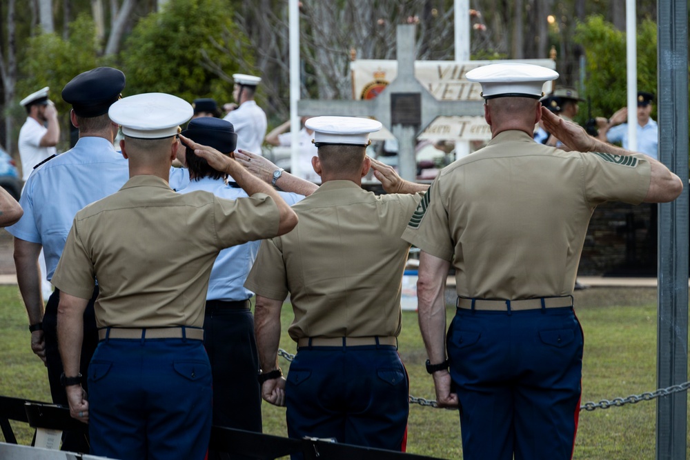 A military ceremony is taking place outdoors, with multiple service members from different branches rendering a salute toward a memorial or monument. Three U.S. Marines in tan MCCUU uniforms and white dress covers are prominently visible from behind, alongside personnel in light blue and white uniforms. A memorial structure with partially visible text including 'VET' is visible in the background.