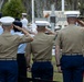 A military ceremony is taking place outdoors, with multiple service members from different branches rendering a salute toward a memorial or monument. Three U.S. Marines in tan MCCUU uniforms and white dress covers are prominently visible from behind, alongside personnel in light blue and white uniforms. A memorial structure with partially visible text including 'VET' is visible in the background.