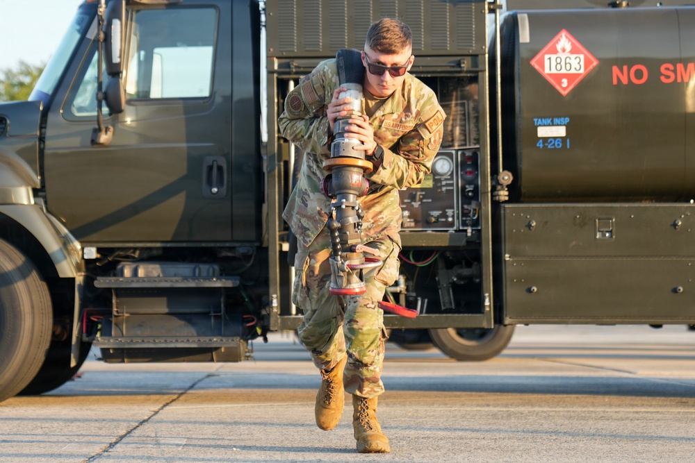 A U.S. Air Force service member in OCP (Operational Camouflage Pattern) uniform is carrying a large fuel nozzle/coupling assembly on a flight line or tarmac. Behind them is a military fuel truck bearing the hazmat placard number 1863, which designates aviation fuel (Jet-A/JP-8). The scene depicts routine aircraft refueling operations at a military airfield.