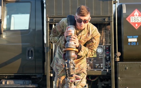 A U.S. Air Force service member in OCP (Operational Camouflage Pattern) uniform is carrying a large fuel nozzle/coupling assembly on a flight line or tarmac. Behind them is a military fuel truck bearing the hazmat placard number 1863, which designates aviation fuel (Jet-A/JP-8). The scene depicts routine aircraft refueling operations at a military airfield.
