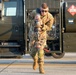 A U.S. Air Force service member in OCP (Operational Camouflage Pattern) uniform is carrying a large fuel nozzle/coupling assembly on a flight line or tarmac. Behind them is a military fuel truck bearing the hazmat placard number 1863, which designates aviation fuel (Jet-A/JP-8). The scene depicts routine aircraft refueling operations at a military airfield.