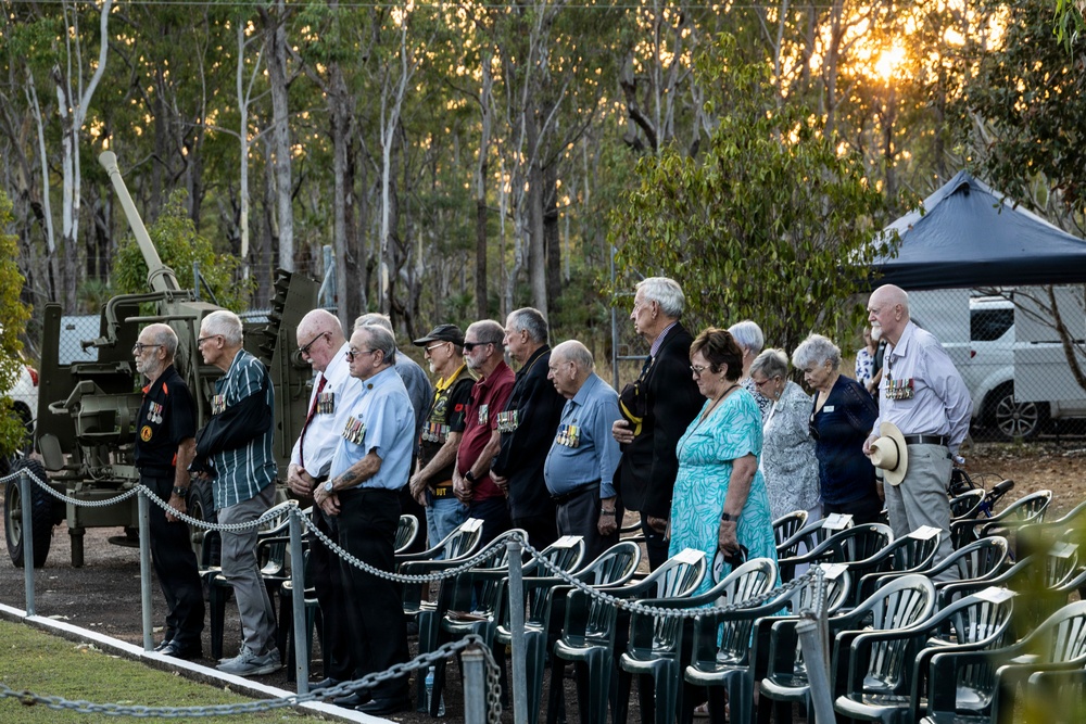 A group of elderly veterans and civilians stand in a line during what appears to be an ANZAC Day or Remembrance Day dawn service in Australia. Several individuals are wearing military service medals on their civilian clothing, and a vintage artillery piece is visible to the left. The ceremony is taking place outdoors at sunrise, surrounded by eucalyptus trees typical of the Australian bush.