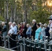 A group of elderly veterans and civilians stand in a line during what appears to be an ANZAC Day or Remembrance Day dawn service in Australia. Several individuals are wearing military service medals on their civilian clothing, and a vintage artillery piece is visible to the left. The ceremony is taking place outdoors at sunrise, surrounded by eucalyptus trees typical of the Australian bush.