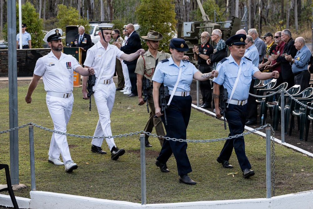 A military ceremonial parade is taking place outdoors, featuring personnel from multiple Australian Defence Force branches including the Royal Australian Navy (white uniforms) and Royal Australian Air Force (light blue shirts, dark navy trousers). The participants are marching in formation carrying rifles, observed by a crowd of civilians and veterans, some wearing medals. The setting appears to be a memorial or commemorative event, likely an ANZAC Day or similar remembrance ceremony.