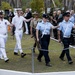 A military ceremonial parade is taking place outdoors, featuring personnel from multiple Australian Defence Force branches including the Royal Australian Navy (white uniforms) and Royal Australian Air Force (light blue shirts, dark navy trousers). The participants are marching in formation carrying rifles, observed by a crowd of civilians and veterans, some wearing medals. The setting appears to be a memorial or commemorative event, likely an ANZAC Day or similar remembrance ceremony.