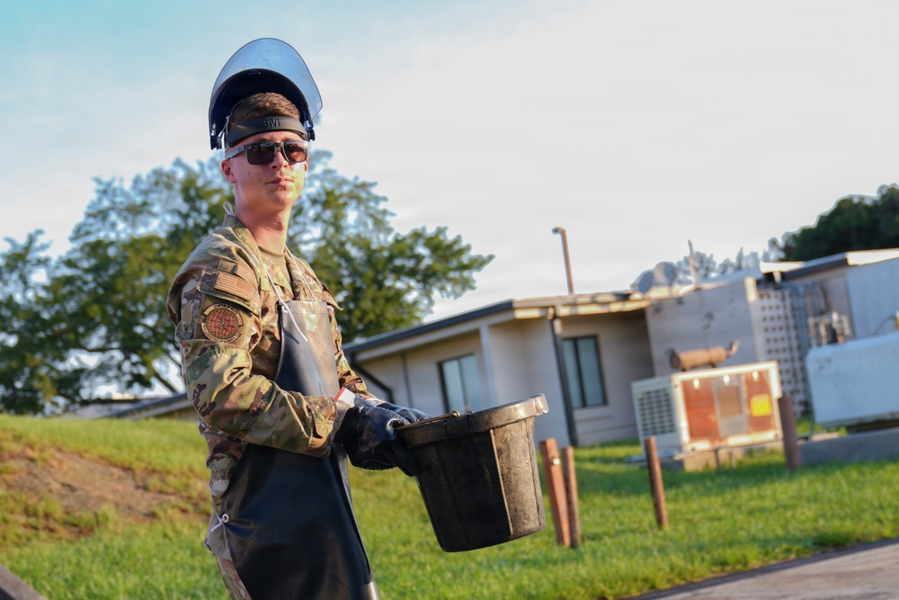 A military service member in camouflage uniform is carrying a dirty bucket while wearing protective gear including a face shield, safety glasses, and black gloves over a rubber apron. The individual appears to be performing a maintenance or cleanup task at what looks like a military installation. The photo is taken outdoors during daylight with buildings and trees visible in the background.