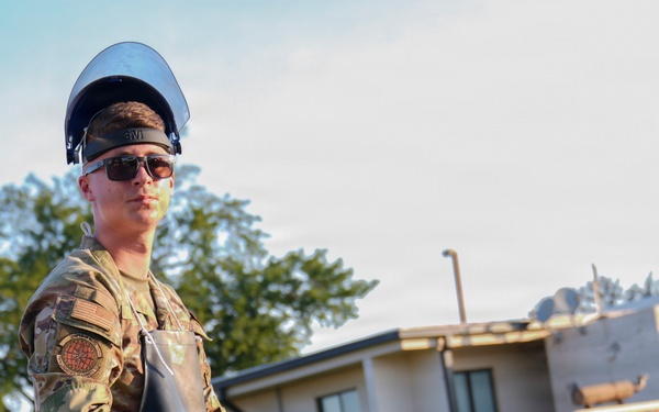 A military service member in camouflage uniform is carrying a dirty bucket while wearing protective gear including a face shield, safety glasses, and black gloves over a rubber apron. The individual appears to be performing a maintenance or cleanup task at what looks like a military installation. The photo is taken outdoors during daylight with buildings and trees visible in the background.