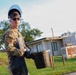 A military service member in camouflage uniform is carrying a dirty bucket while wearing protective gear including a face shield, safety glasses, and black gloves over a rubber apron. The individual appears to be performing a maintenance or cleanup task at what looks like a military installation. The photo is taken outdoors during daylight with buildings and trees visible in the background.