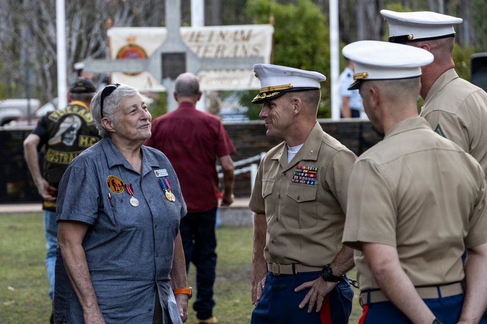 A civilian woman wearing medals and patches is engaged in conversation with U.S. Marine Corps officers in service uniforms at what appears to be a Vietnam Veterans memorial event. The setting is an outdoor memorial or park area with a banner reading 'Vietnam Veterans' visible in the background. The gathering appears to be a commemorative ceremony or reunion event.