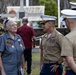 A civilian woman wearing medals and patches is engaged in conversation with U.S. Marine Corps officers in service uniforms at what appears to be a Vietnam Veterans memorial event. The setting is an outdoor memorial or park area with a banner reading 'Vietnam Veterans' visible in the background. The gathering appears to be a commemorative ceremony or reunion event.