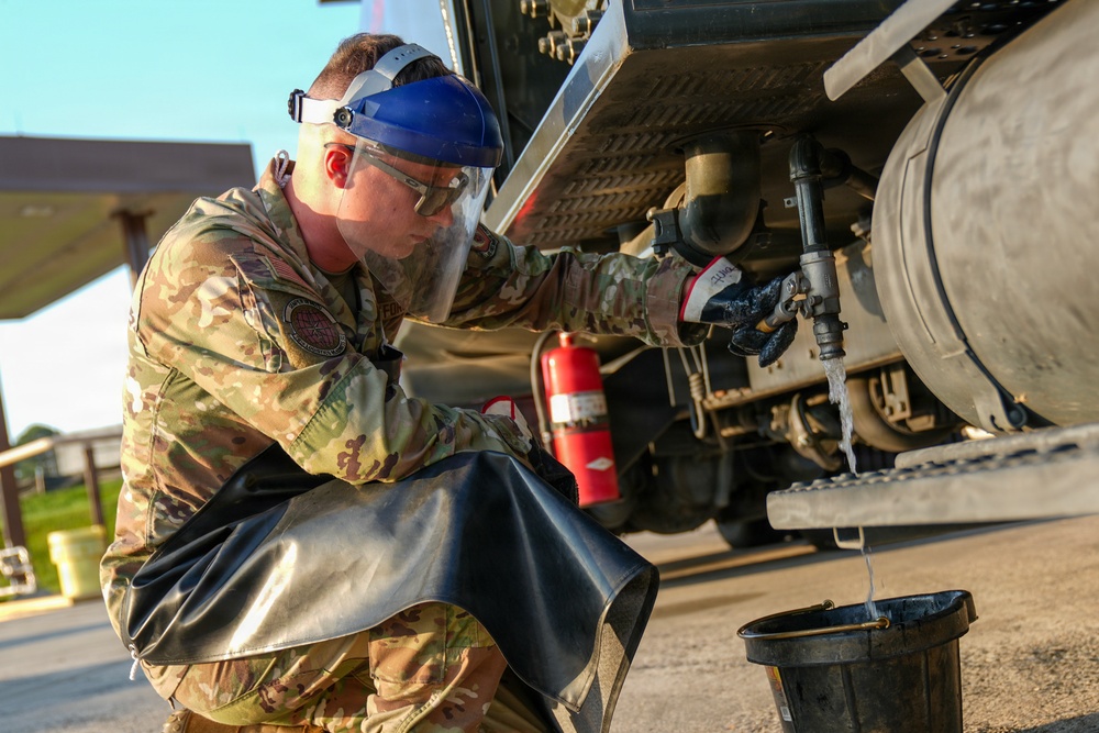 A military service member in US Air Force OCP (Operational Camouflage Pattern) uniform is performing maintenance or inspection work on a fuel or liquid tanker vehicle. The individual is wearing full personal protective equipment including a face shield, safety glasses, and gloves while draining or sampling liquid into a black bucket. A red fire extinguisher is visible mounted beneath the vehicle, indicating standard safety protocols are in place.
