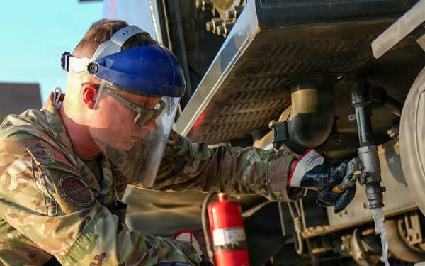 A military service member in US Air Force OCP (Operational Camouflage Pattern) uniform is performing maintenance or inspection work on a fuel or liquid tanker vehicle. The individual is wearing full personal protective equipment including a face shield, safety glasses, and gloves while draining or sampling liquid into a black bucket. A red fire extinguisher is visible mounted beneath the vehicle, indicating standard safety protocols are in place.
