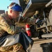 A military service member in US Air Force OCP (Operational Camouflage Pattern) uniform is performing maintenance or inspection work on a fuel or liquid tanker vehicle. The individual is wearing full personal protective equipment including a face shield, safety glasses, and gloves while draining or sampling liquid into a black bucket. A red fire extinguisher is visible mounted beneath the vehicle, indicating standard safety protocols are in place.