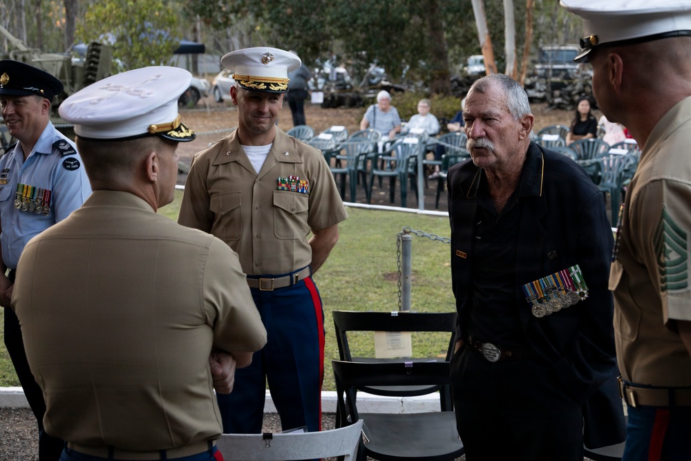 A military ceremony or informal gathering featuring U.S. Marine Corps personnel in service uniforms interacting with a civilian veteran wearing service medals on a black shirt. An Australian Air Force officer is also present on the left, identifiable by his light blue uniform and medals. The setting appears to be an outdoor military museum or memorial site, with military vehicles and equipment visible in the background.