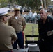 A military ceremony or informal gathering featuring U.S. Marine Corps personnel in service uniforms interacting with a civilian veteran wearing service medals on a black shirt. An Australian Air Force officer is also present on the left, identifiable by his light blue uniform and medals. The setting appears to be an outdoor military museum or memorial site, with military vehicles and equipment visible in the background.