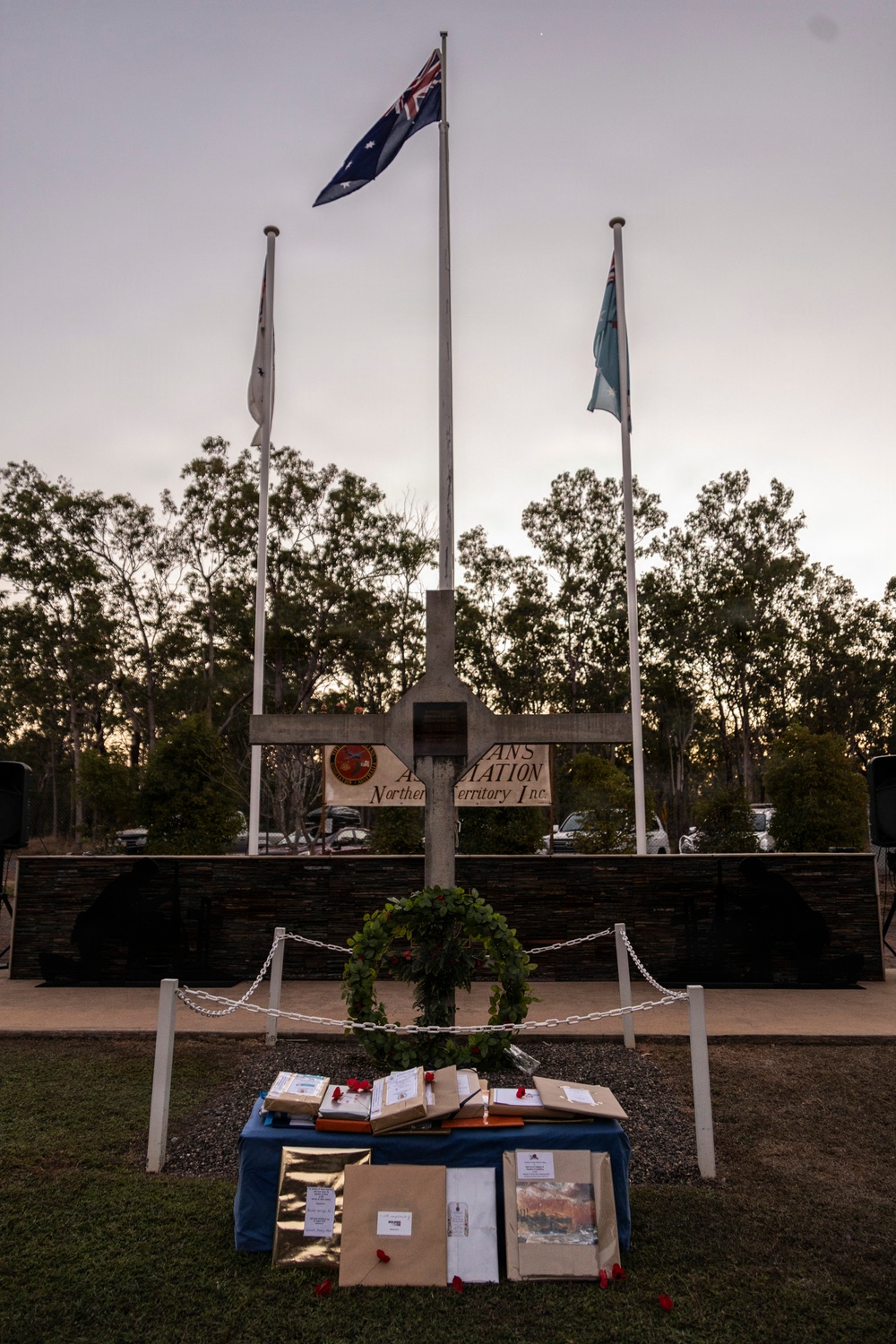 A memorial ceremony setting at what appears to be a Veterans Association Northern Territory Inc. memorial site, photographed at dusk or dawn. A large stone cross serves as the central monument, flanked by three flagpoles including the Australian national flag. A table covered with a blue cloth holds wrapped packages and items adorned with red poppies, positioned in front of a wreath-decorated cross.
