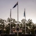 A memorial ceremony setting at what appears to be a Veterans Association Northern Territory Inc. memorial site, photographed at dusk or dawn. A large stone cross serves as the central monument, flanked by three flagpoles including the Australian national flag. A table covered with a blue cloth holds wrapped packages and items adorned with red poppies, positioned in front of a wreath-decorated cross.