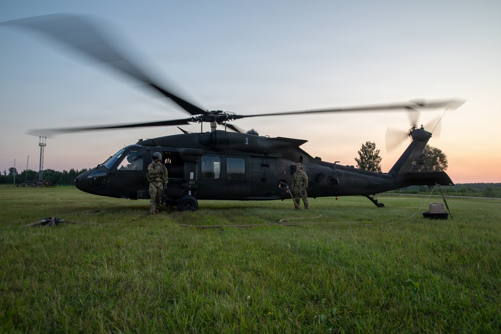 A dark-colored UH-60 Black Hawk helicopter marked 'United States Army' is on the ground in a grassy field during dusk or dawn conditions. Two soldiers in military fatigues are standing near the aircraft, which has its rotors spinning. Ground support equipment is visible alongside the helicopter.