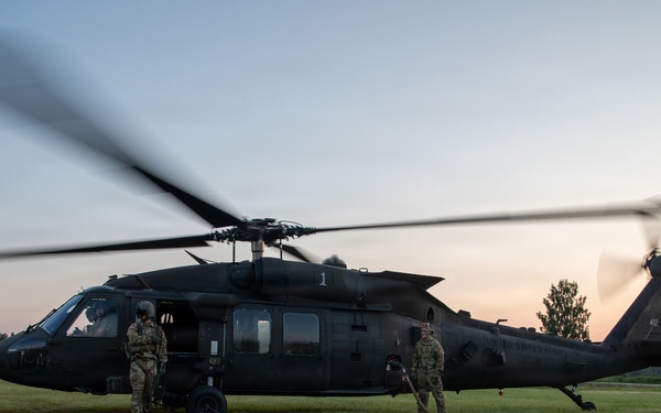 A dark-colored UH-60 Black Hawk helicopter marked 'United States Army' is on the ground in a grassy field during dusk or dawn conditions. Two soldiers in military fatigues are standing near the aircraft, which has its rotors spinning. Ground support equipment is visible alongside the helicopter.