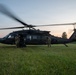 A dark-colored UH-60 Black Hawk helicopter marked 'United States Army' is on the ground in a grassy field during dusk or dawn conditions. Two soldiers in military fatigues are standing near the aircraft, which has its rotors spinning. Ground support equipment is visible alongside the helicopter.