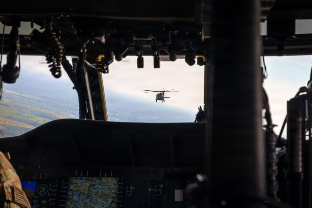 The image is taken from inside a military helicopter cockpit, looking forward through the windshield at another helicopter flying in formation ahead. The interior cockpit instrumentation, including a digital map display, is visible in the foreground. The terrain below appears to be flat agricultural land viewed from altitude.