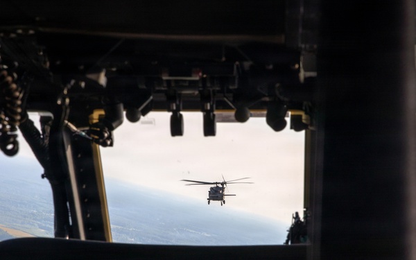 The image is taken from inside a military helicopter cockpit, looking forward through the windshield at another helicopter flying in formation ahead. The interior cockpit instrumentation, including a digital map display, is visible in the foreground. The terrain below appears to be flat agricultural land viewed from altitude.