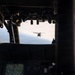 The image is taken from inside a military helicopter cockpit, looking forward through the windshield at another helicopter flying in formation ahead. The interior cockpit instrumentation, including a digital map display, is visible in the foreground. The terrain below appears to be flat agricultural land viewed from altitude.