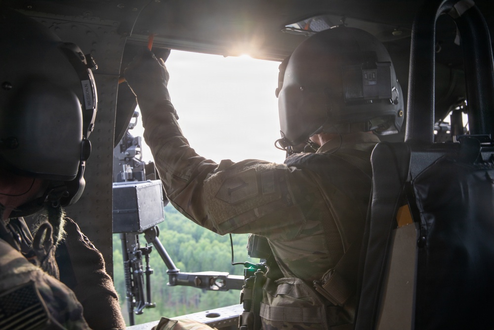 Two military personnel are seated inside a helicopter in flight, with one individual reaching upward toward the window frame or overhead panel. The interior shows tactical equipment including a mounted weapon system visible through the open side door. Green forested terrain is visible below through the open door, suggesting the aircraft is airborne at low-to-medium altitude.