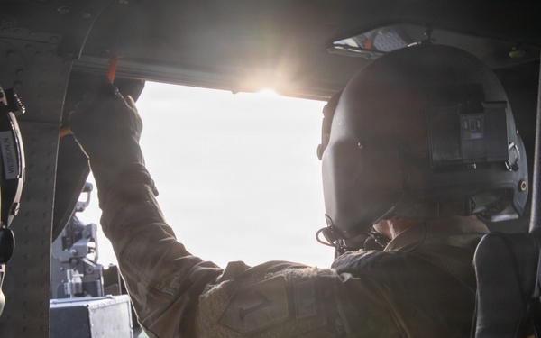 Two military personnel are seated inside a helicopter in flight, with one individual reaching upward toward the window frame or overhead panel. The interior shows tactical equipment including a mounted weapon system visible through the open side door. Green forested terrain is visible below through the open door, suggesting the aircraft is airborne at low-to-medium altitude.