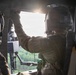 Two military personnel are seated inside a helicopter in flight, with one individual reaching upward toward the window frame or overhead panel. The interior shows tactical equipment including a mounted weapon system visible through the open side door. Green forested terrain is visible below through the open door, suggesting the aircraft is airborne at low-to-medium altitude.