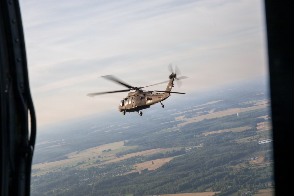 A military helicopter, consistent with a UH-60 Black Hawk, is photographed in flight from the open door of another aircraft. The image is taken at altitude, revealing a broad agricultural landscape with fields, forests, and scattered settlements below. The photo appears to be taken during a formation or escort flight.