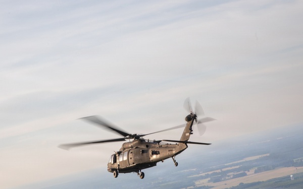 A military helicopter, consistent with a UH-60 Black Hawk, is photographed in flight from the open door of another aircraft. The image is taken at altitude, revealing a broad agricultural landscape with fields, forests, and scattered settlements below. The photo appears to be taken during a formation or escort flight.