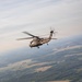 A military helicopter, consistent with a UH-60 Black Hawk, is photographed in flight from the open door of another aircraft. The image is taken at altitude, revealing a broad agricultural landscape with fields, forests, and scattered settlements below. The photo appears to be taken during a formation or escort flight.