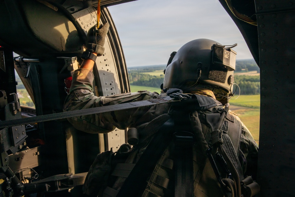 A military aircrew member is photographed from behind while seated inside a helicopter in flight. The individual is wearing full tactical gear including a flight helmet with mounted equipment, multicam uniform, and tactical vest. The open door of the helicopter reveals a rural landscape with green fields and tree lines below.