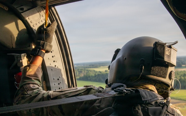 A military aircrew member is photographed from behind while seated inside a helicopter in flight. The individual is wearing full tactical gear including a flight helmet with mounted equipment, multicam uniform, and tactical vest. The open door of the helicopter reveals a rural landscape with green fields and tree lines below.