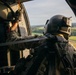 A military aircrew member is photographed from behind while seated inside a helicopter in flight. The individual is wearing full tactical gear including a flight helmet with mounted equipment, multicam uniform, and tactical vest. The open door of the helicopter reveals a rural landscape with green fields and tree lines below.