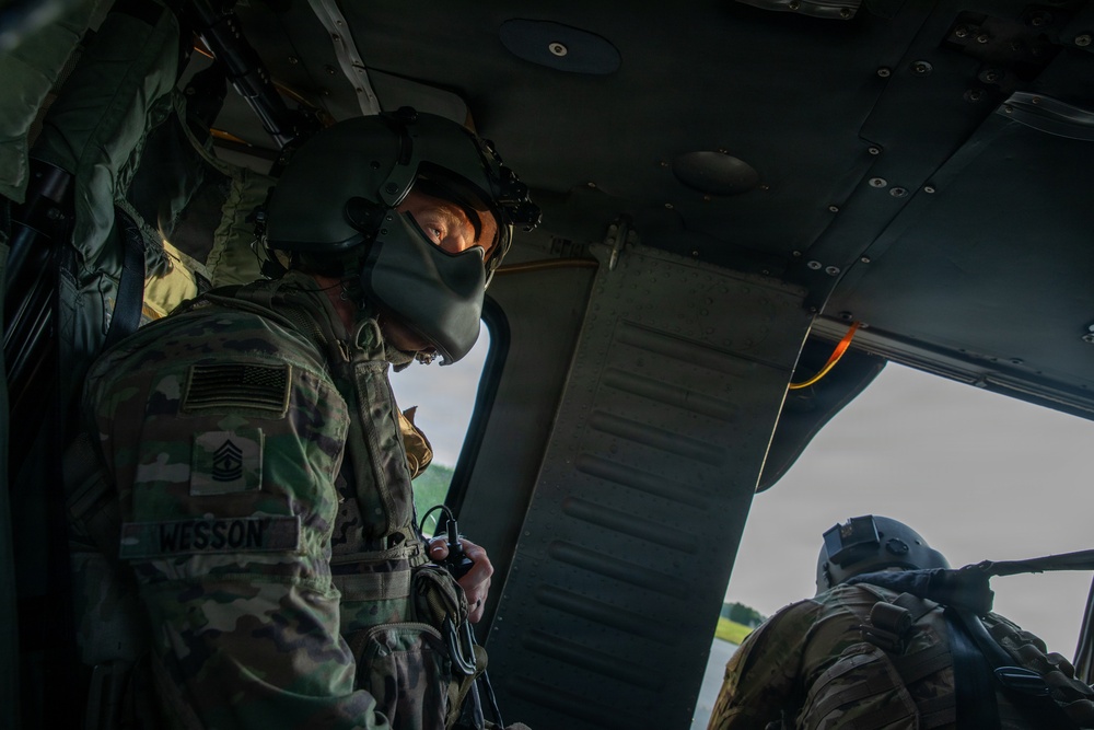 Two U.S. Army soldiers are photographed inside a military helicopter, likely a UH-60 Black Hawk. The soldier in the foreground, identified by name tape as 'WESSON' with a Master Sergeant rank insignia, is wearing a flight helmet and MultiCam (OCP) uniform with a U.S. flag patch. A second soldier is visible in the background near an open door, appearing to man a door gun position.