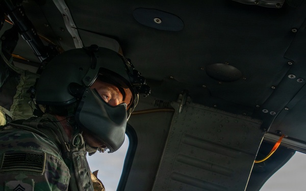 Two U.S. Army soldiers are photographed inside a military helicopter, likely a UH-60 Black Hawk. The soldier in the foreground, identified by name tape as 'WESSON' with a Master Sergeant rank insignia, is wearing a flight helmet and MultiCam (OCP) uniform with a U.S. flag patch. A second soldier is visible in the background near an open door, appearing to man a door gun position.