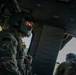 Two U.S. Army soldiers are photographed inside a military helicopter, likely a UH-60 Black Hawk. The soldier in the foreground, identified by name tape as 'WESSON' with a Master Sergeant rank insignia, is wearing a flight helmet and MultiCam (OCP) uniform with a U.S. flag patch. A second soldier is visible in the background near an open door, appearing to man a door gun position.