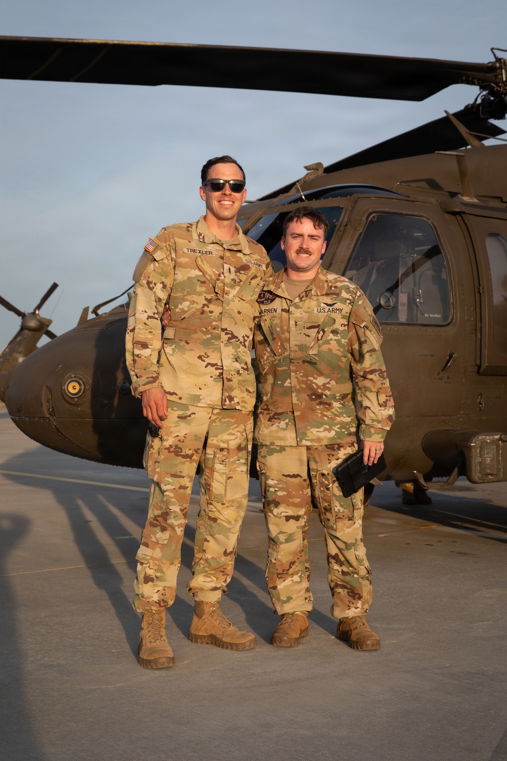 Two U.S. Army soldiers in Operational Camouflage Pattern (OCP) uniforms are posing together on a flight line in front of a UH-60 Black Hawk helicopter. Name tapes visible on their uniforms read 'TREXLER' (left) and 'WARREN' (right), both bearing 'U.S. ARMY' tape. The photo appears to be taken during golden hour lighting at a military airfield.