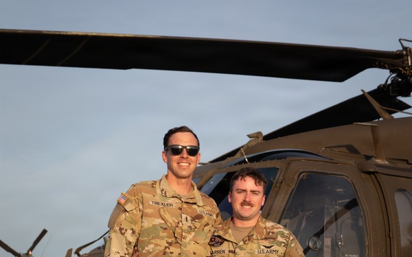 Two U.S. Army soldiers in Operational Camouflage Pattern (OCP) uniforms are posing together on a flight line in front of a UH-60 Black Hawk helicopter. Name tapes visible on their uniforms read 'TREXLER' (left) and 'WARREN' (right), both bearing 'U.S. ARMY' tape. The photo appears to be taken during golden hour lighting at a military airfield.