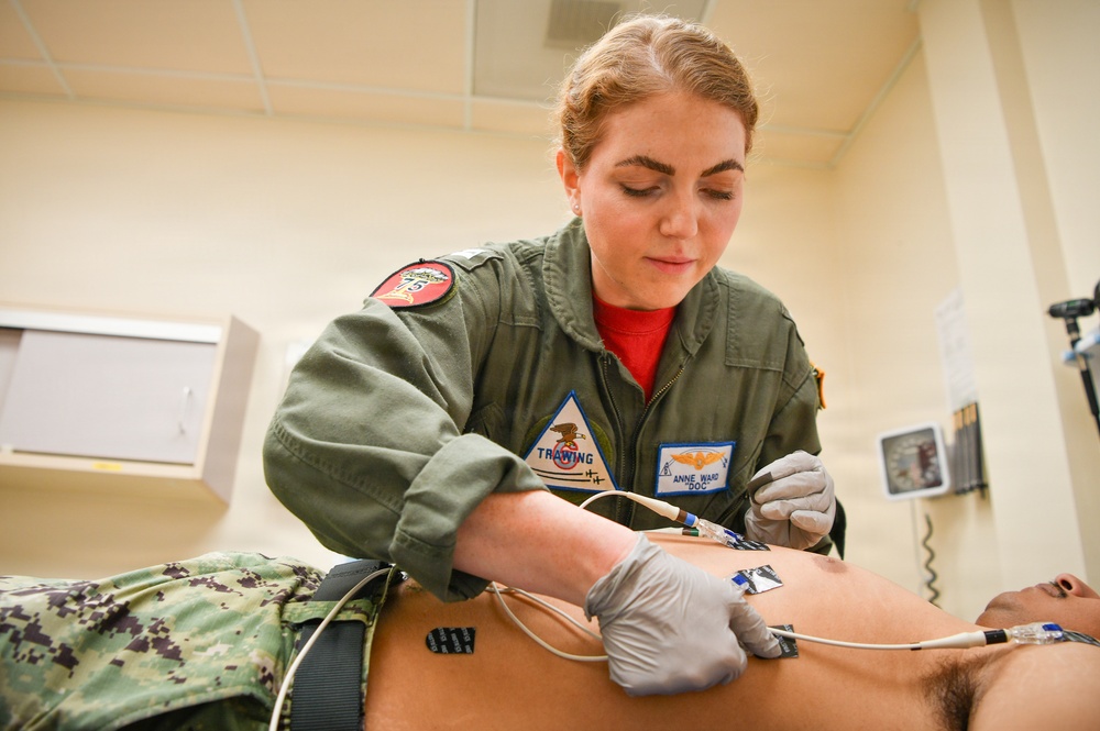 A military medical professional in an olive green flight suit is performing a medical procedure on a patient lying on an examination table. The provider, identified by name tag as 'Anne Ward' with the callsign 'Doc', is wearing latex gloves and attaching ECG/EKG electrodes or medical leads to the patient's torso. The setting appears to be a military medical facility or sick bay.
