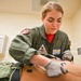 A military medical professional in an olive green flight suit is performing a medical procedure on a patient lying on an examination table. The provider, identified by name tag as 'Anne Ward' with the callsign 'Doc', is wearing latex gloves and attaching ECG/EKG electrodes or medical leads to the patient's torso. The setting appears to be a military medical facility or sick bay.