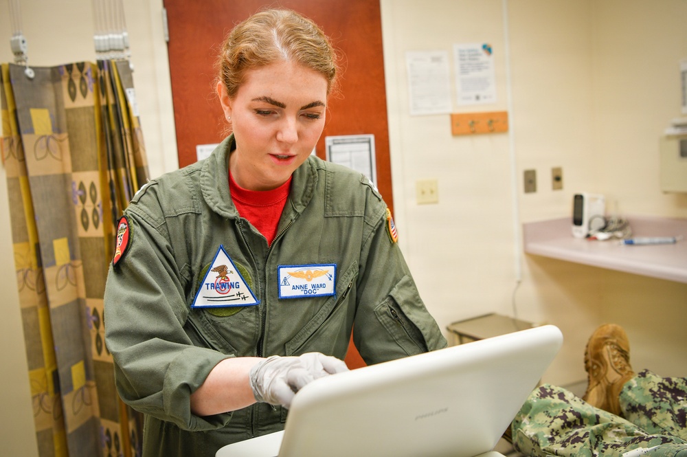 A female military medical personnel wearing an olive green flight suit is working on a white laptop while wearing latex gloves. Her name tag reads 'ANNE WARD' with the nickname 'DOC', and she has a 'TRAINING' patch on her chest along with a flight surgeon wings insignia. The setting appears to be a medical examination room or sick bay facility.