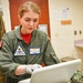 A female military medical personnel wearing an olive green flight suit is working on a white laptop while wearing latex gloves. Her name tag reads 'ANNE WARD' with the nickname 'DOC', and she has a 'TRAINING' patch on her chest along with a flight surgeon wings insignia. The setting appears to be a medical examination room or sick bay facility.