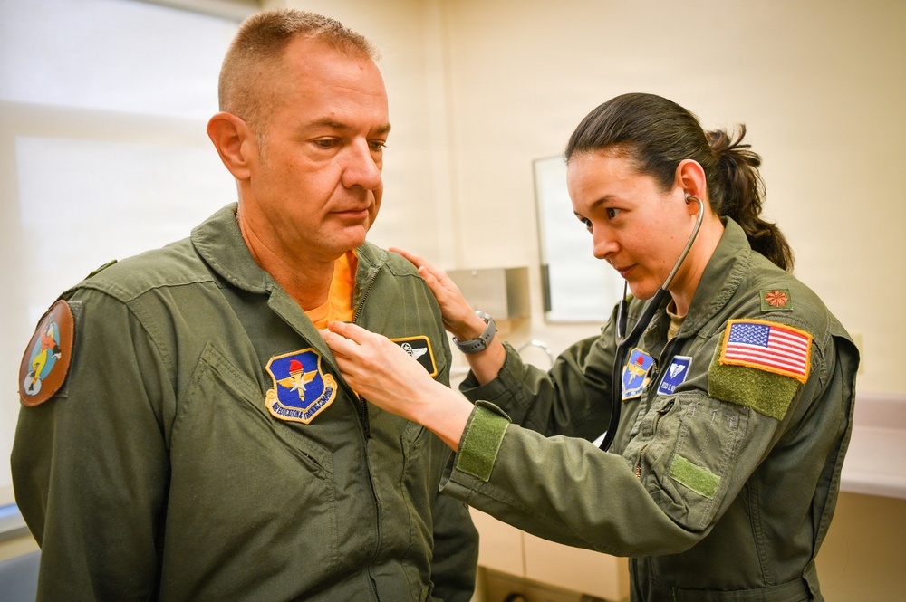 Two individuals wearing olive green military flight suits are in what appears to be a medical examination room. A female military medical personnel is using a stethoscope to examine a male military member's chest area. Both wear unit patches and the female has a US flag patch on her right sleeve.