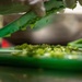 A close-up shot of someone chopping green herbs or vegetables on a green cutting board. The image is taken from a very low angle, showing finely minced greens being scraped with a green knife. The shallow depth of field creates a blurred background with what appears to be a commercial kitchen setting.