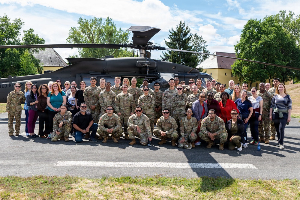 A large group of approximately 50 individuals, consisting of both military personnel in camouflage uniforms and civilians in casual attire, pose for a group photograph in front of a United States Army UH-60 Black Hawk helicopter. The photo appears to be taken on a military installation or airfield on a sunny day with partial cloud cover. The setting includes green trees and what appear to be military or institutional buildings in the background.