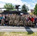 A large group of approximately 50 individuals, consisting of both military personnel in camouflage uniforms and civilians in casual attire, pose for a group photograph in front of a United States Army UH-60 Black Hawk helicopter. The photo appears to be taken on a military installation or airfield on a sunny day with partial cloud cover. The setting includes green trees and what appear to be military or institutional buildings in the background.